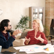 Young bearded designer and his happy mature female colleague sitting by table with many papers, brainstorming and discussing new ideas using body language