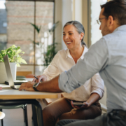 A man and a woman with a laptop, meeting in an office, are negotiating cooperatively, the woman is smiling.