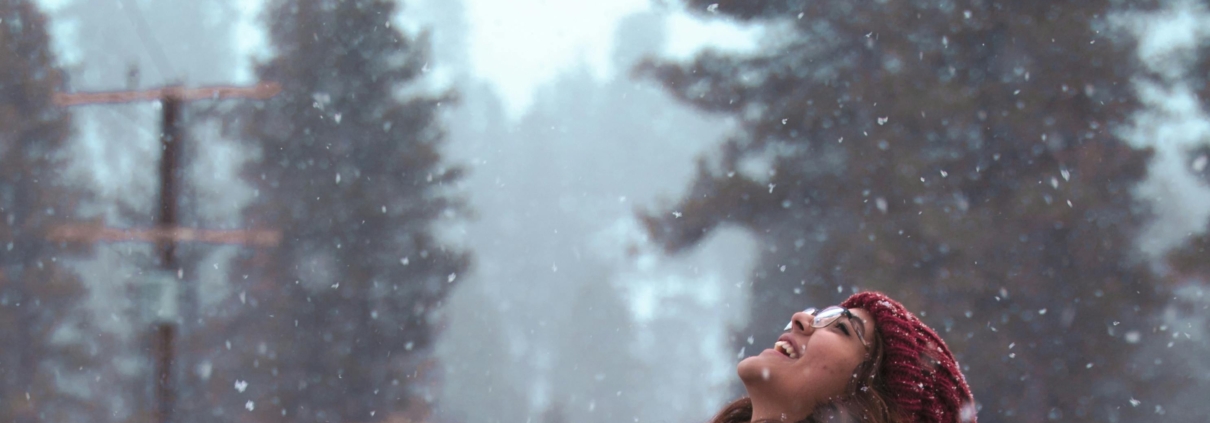 woman standing with her head up watching the snow, wearing a woolly red hat and a grey coat