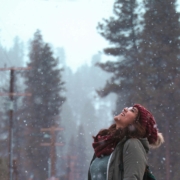 woman standing with her head up watching the snow, wearing a woolly red hat and a grey coat