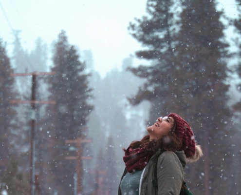 woman standing with her head up watching the snow, wearing a woolly red hat and a grey coat