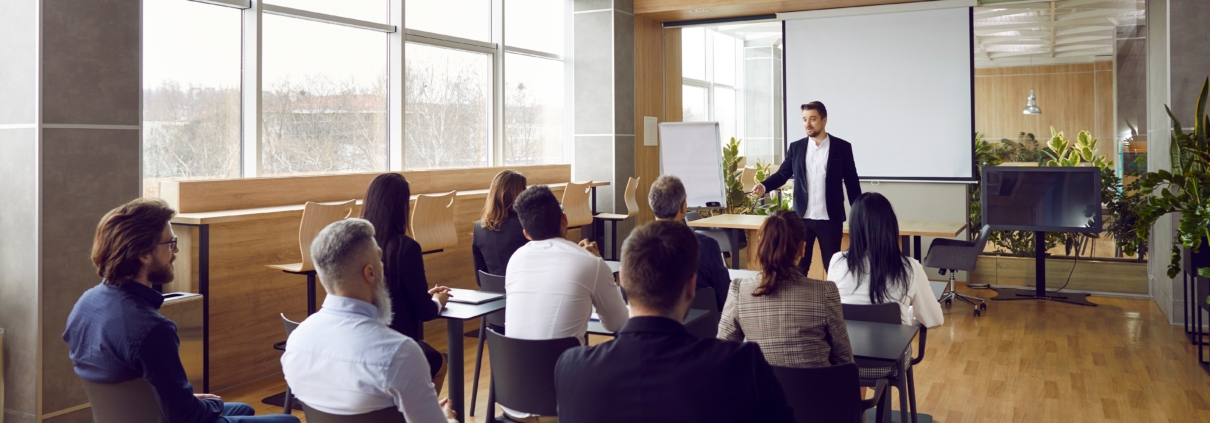 In a modern, sunlit conference room, a confident presenter engages a diverse audience. The setting features a projector screen, varied seating, and a décor of wood and greenery, promoting warmth and creativity. The session, titled "Communication Confidence: Presentation Strategies" highlights effective communication and public speaking.