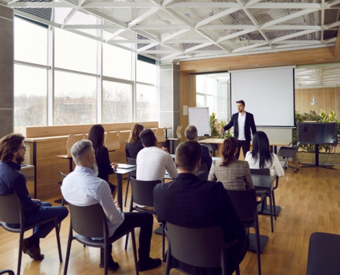 In a modern, sunlit conference room, a confident presenter engages a diverse audience. The setting features a projector screen, varied seating, and a décor of wood and greenery, promoting warmth and creativity. The session, titled "Communication Confidence: Presentation Strategies" highlights effective communication and public speaking.