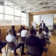 In a modern, sunlit conference room, a confident presenter engages a diverse audience. The setting features a projector screen, varied seating, and a décor of wood and greenery, promoting warmth and creativity. The session, titled "Communication Confidence: Presentation Strategies" highlights effective communication and public speaking.