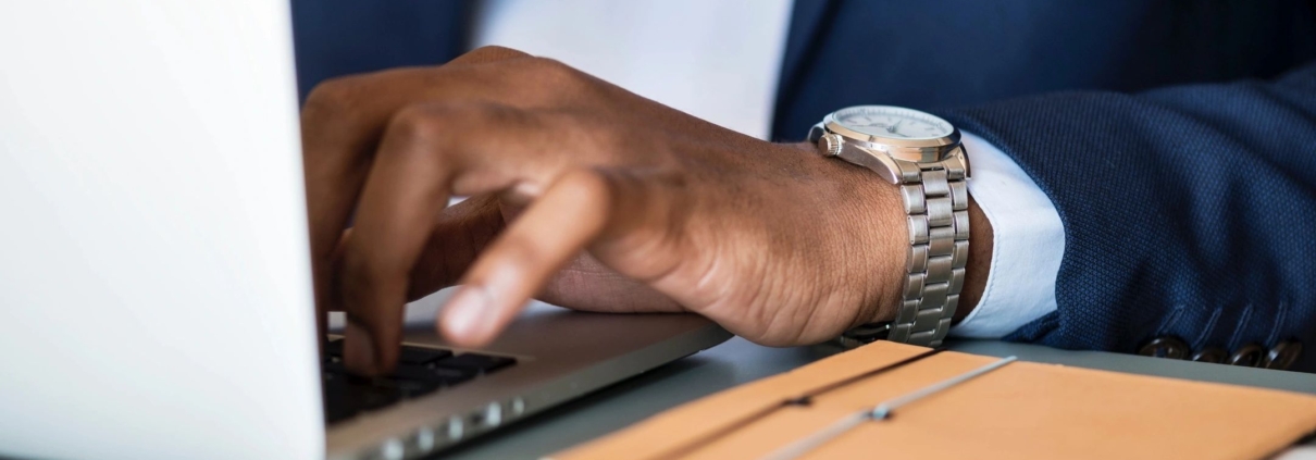 A man's hand is rested on a laptop keyboard, his watch is showing out of his light blue sleeve, and an orange notebook is on the table next to him