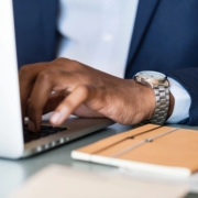 A man's hand is rested on a laptop keyboard, his watch is showing out of his light blue sleeve, and an orange notebook is on the table next to him