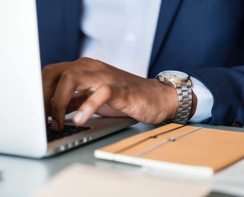 A man's hand is rested on a laptop keyboard, his watch is showing out of his light blue sleeve, and an orange notebook is on the table next to him