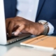 A man's hand is rested on a laptop keyboard, his watch is showing out of his light blue sleeve, and an orange notebook is on the table next to him