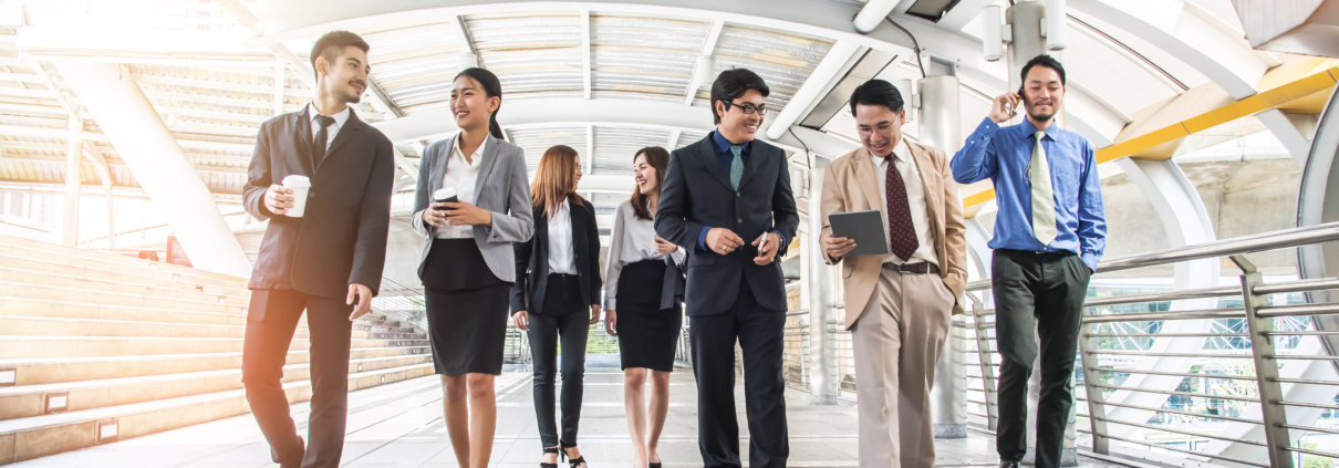 A group of business professionals walking together through a modern, covered walkway, holding laptops, notebooks, and coffee cups, symbolising teamwork and leadership in a corporate environment.