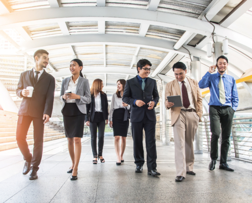 A group of business professionals walking together through a modern, covered walkway, holding laptops, notebooks, and coffee cups, symbolising teamwork and leadership in a corporate environment.